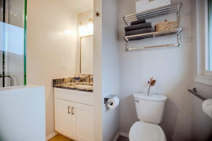 Bathroom with white vanity, granite countertop, mirror, toilet, towel shelf, and window with blinds