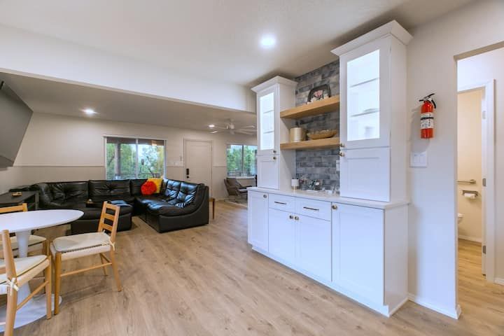 Bright open kitchen-living area with white cabinets, wooden floor, dining table, and black sofa by windows