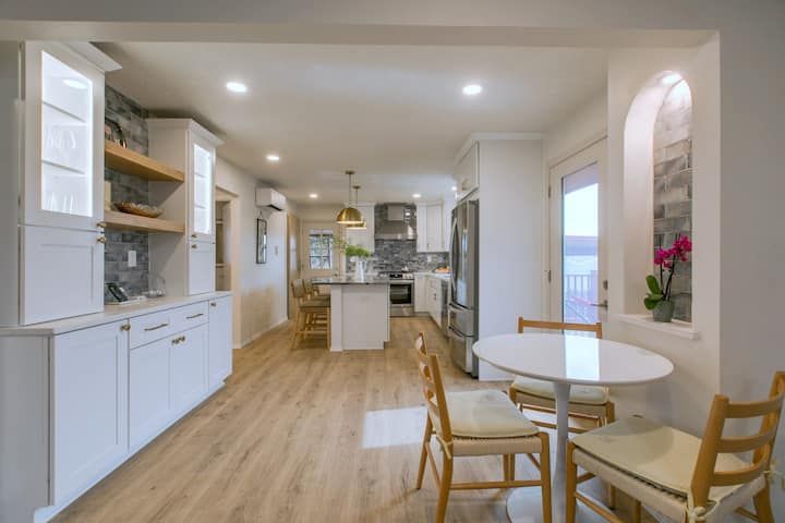 Bright white kitchen with island, wood floors, and a small round dining table by an arched wall niche.