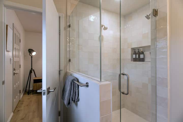 Modern glass shower with beige tile and an open door to a bedroom with a floor lamp.