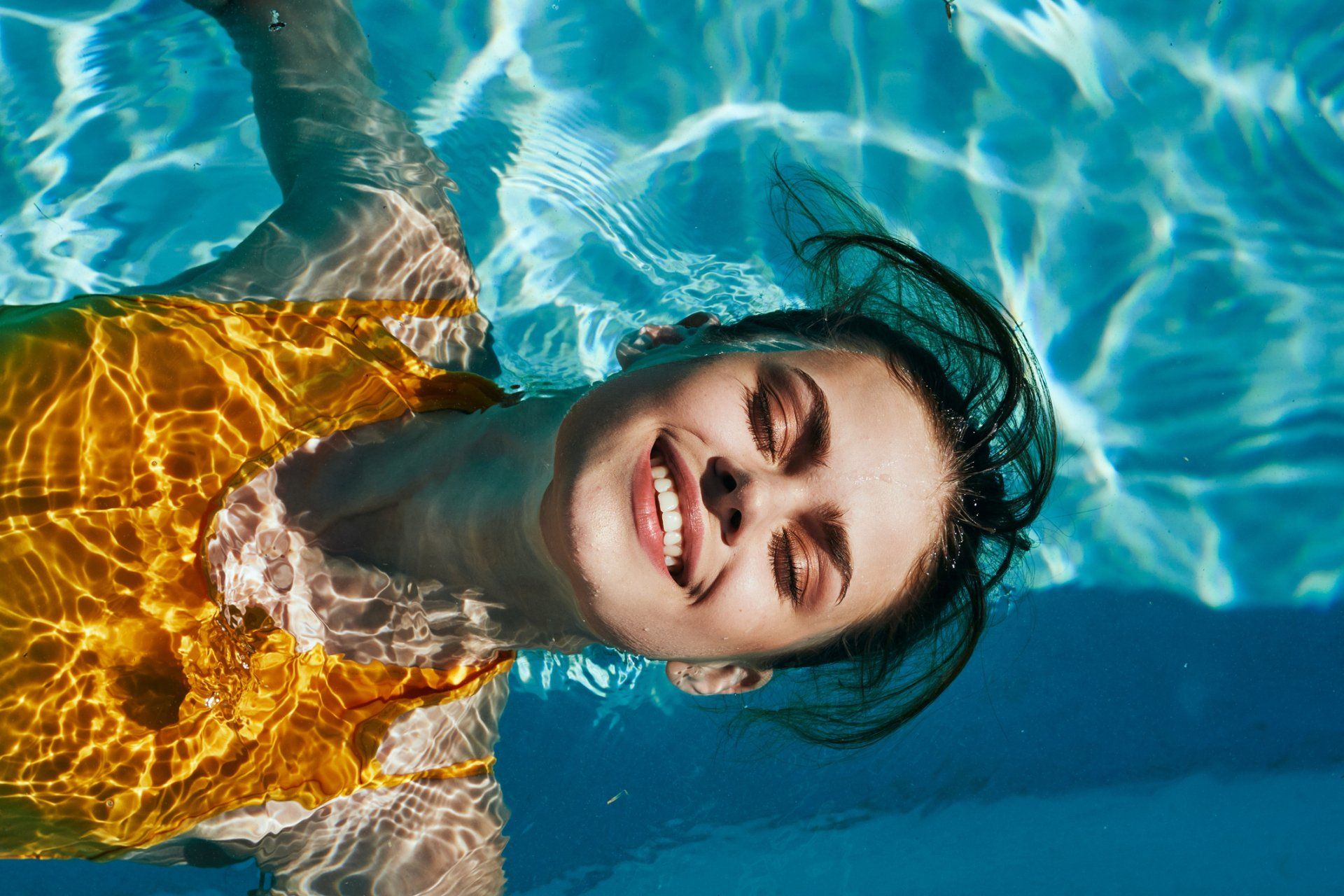 Woman smiling underwater in pool, wearing yellow top.