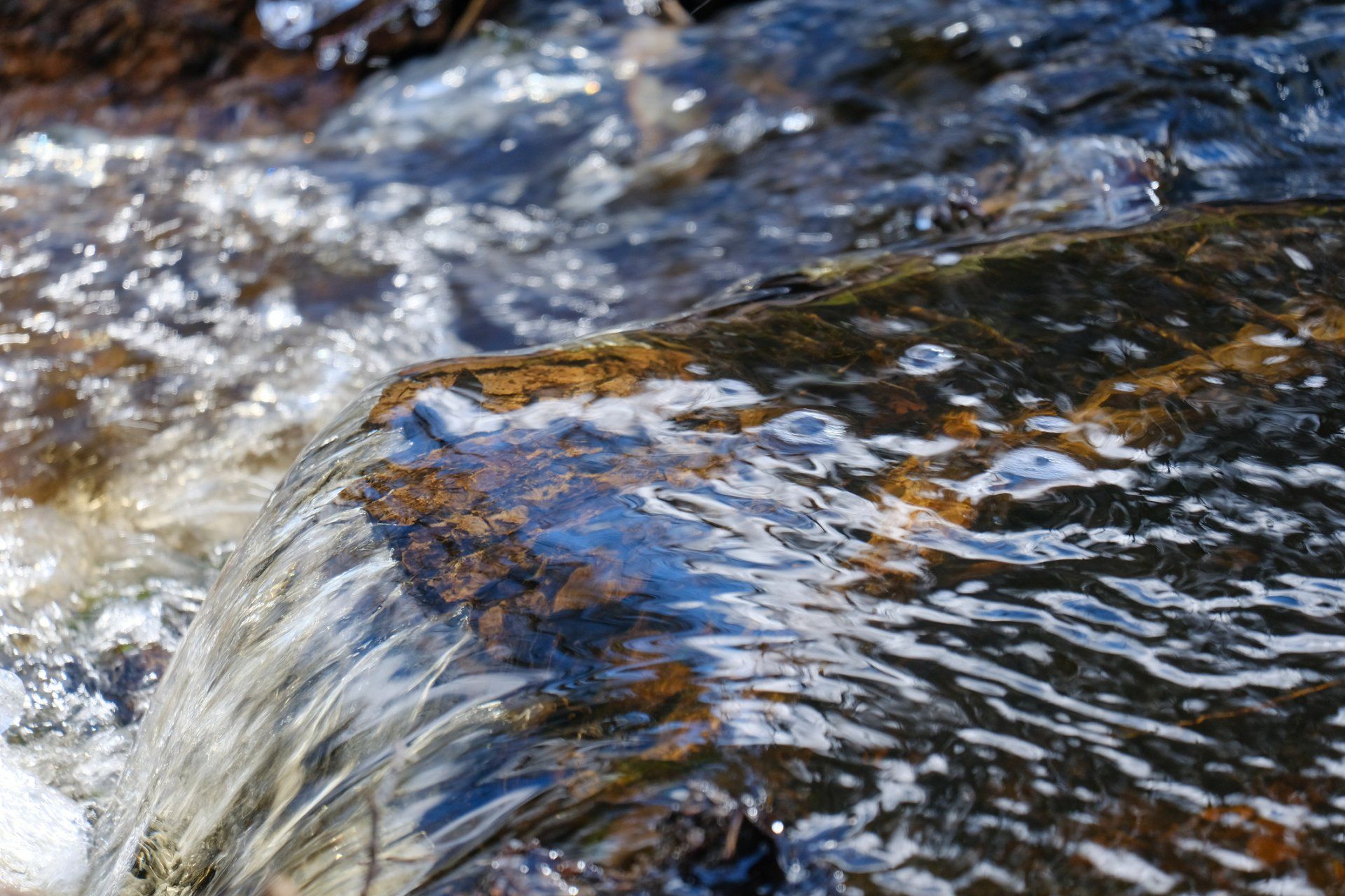 Water Flowing — Muncie, IN — Oxley Softwater Co. Close-up of water cascading over a dark, textured rock in a stream. Sunlight glistens on the water.