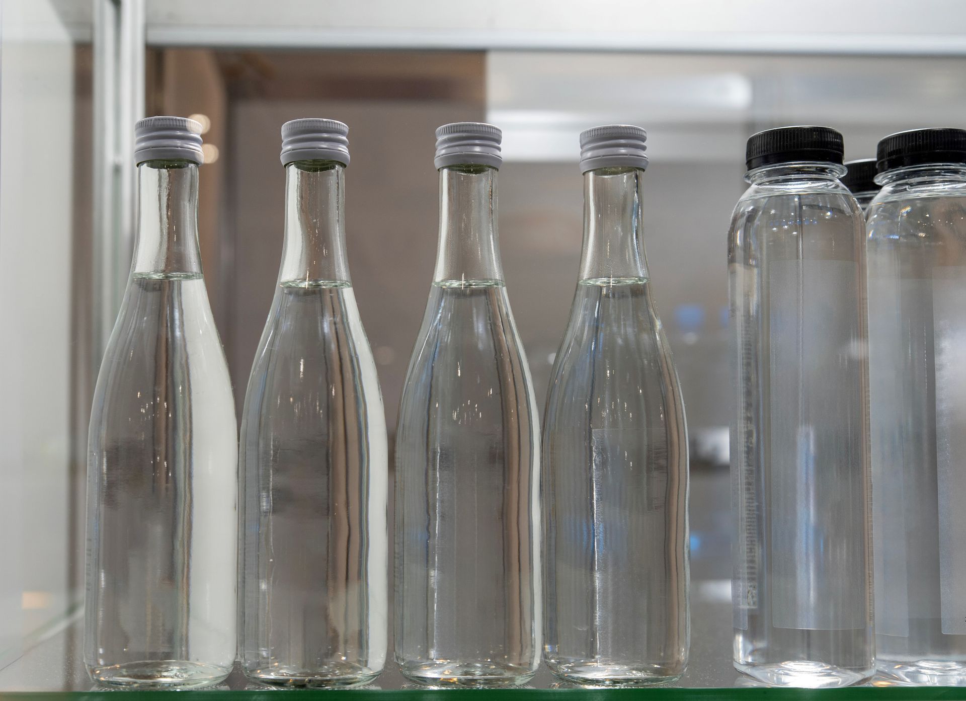 Glass water bottles with silver lids. Bottles of water in glass and plastic with silver and black caps on a shelf.
