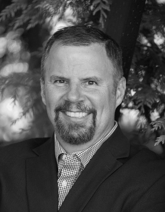 Portrait of a smiling man in a suit and checkered shirt against a blurred outdoor background