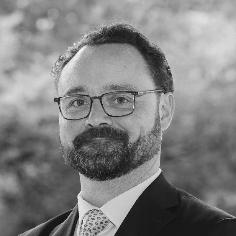 Black-and-white headshot of a bearded man wearing glasses, a suit, and a tie, smiling slightly.
