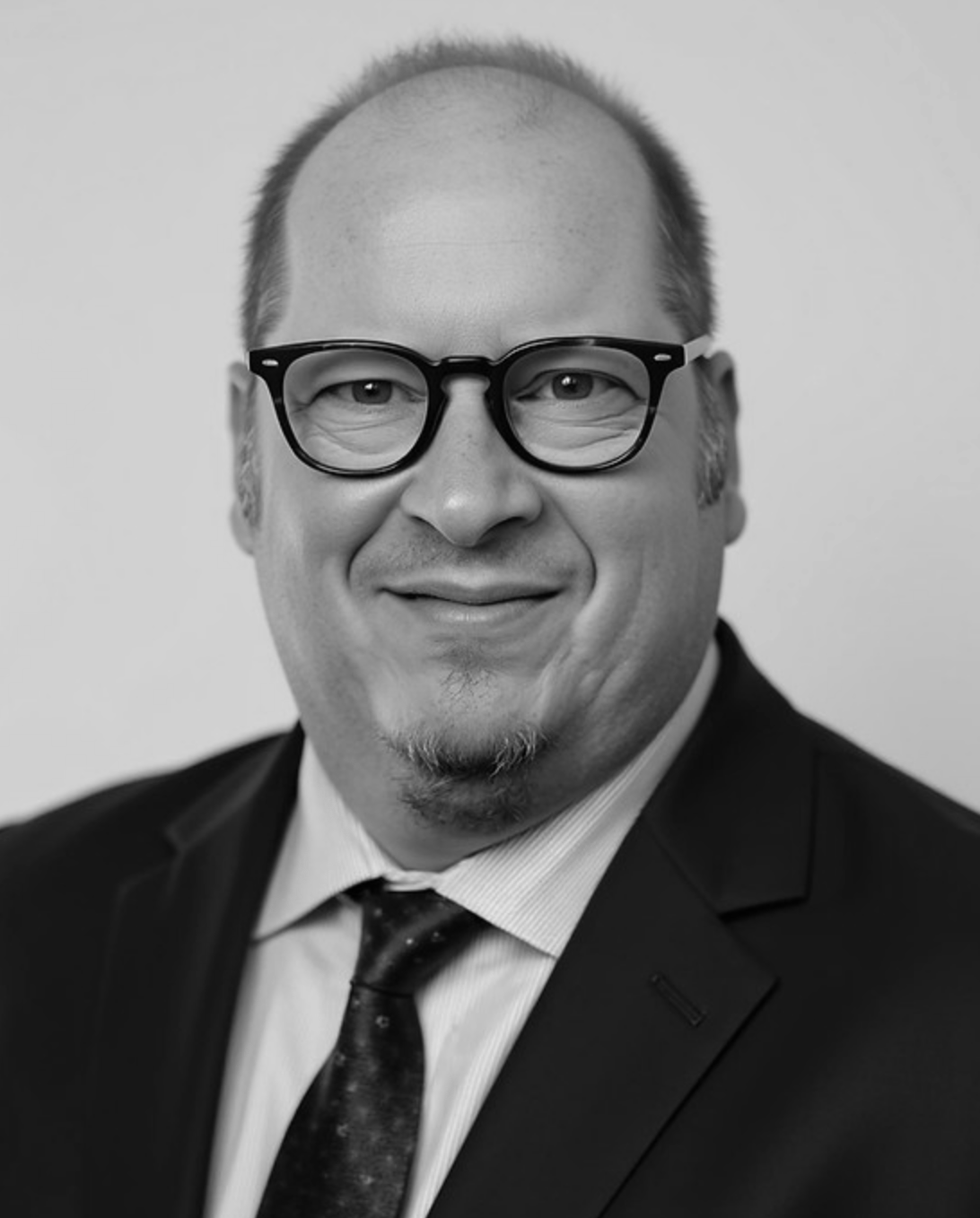 Headshot of a man in glasses and a suit, smiling against a plain background
