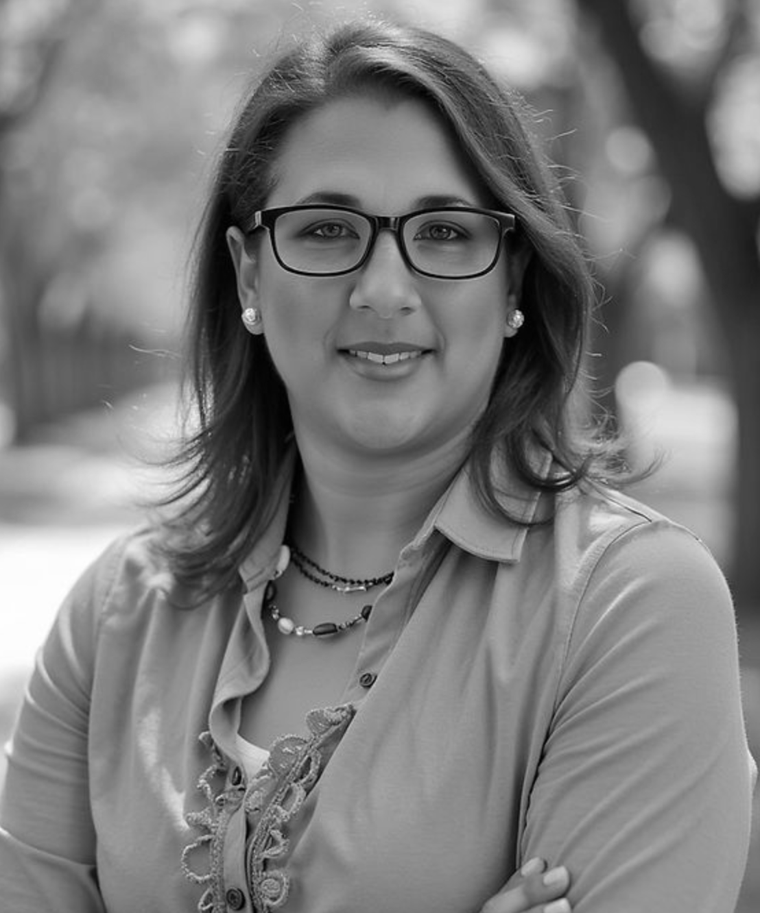 Smiling person wearing glasses and a collared shirt, posed outdoors with a blurred background.
