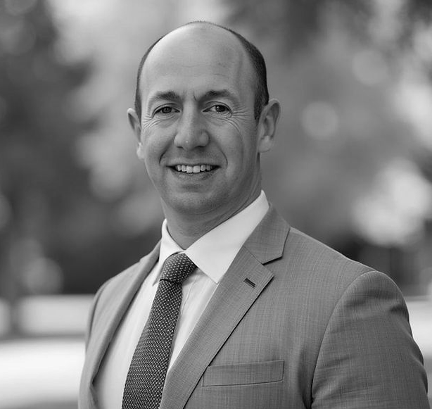 Smiling man in a suit and tie outdoors, with a blurred background