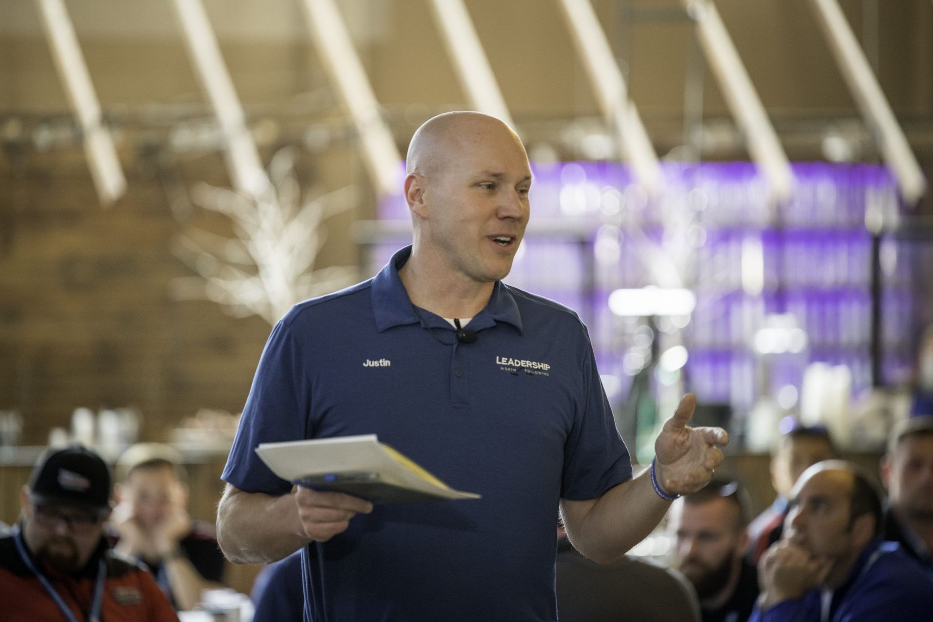 A man in a blue shirt is holding a clipboard and talking to a group of people.