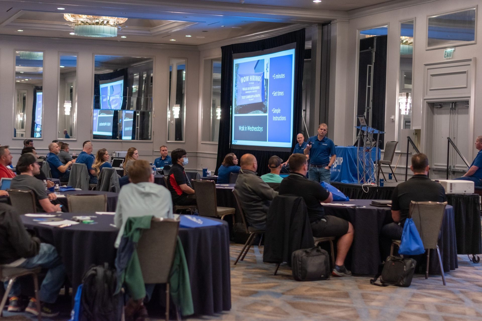 A group of people are sitting at tables in a conference room watching a presentation.