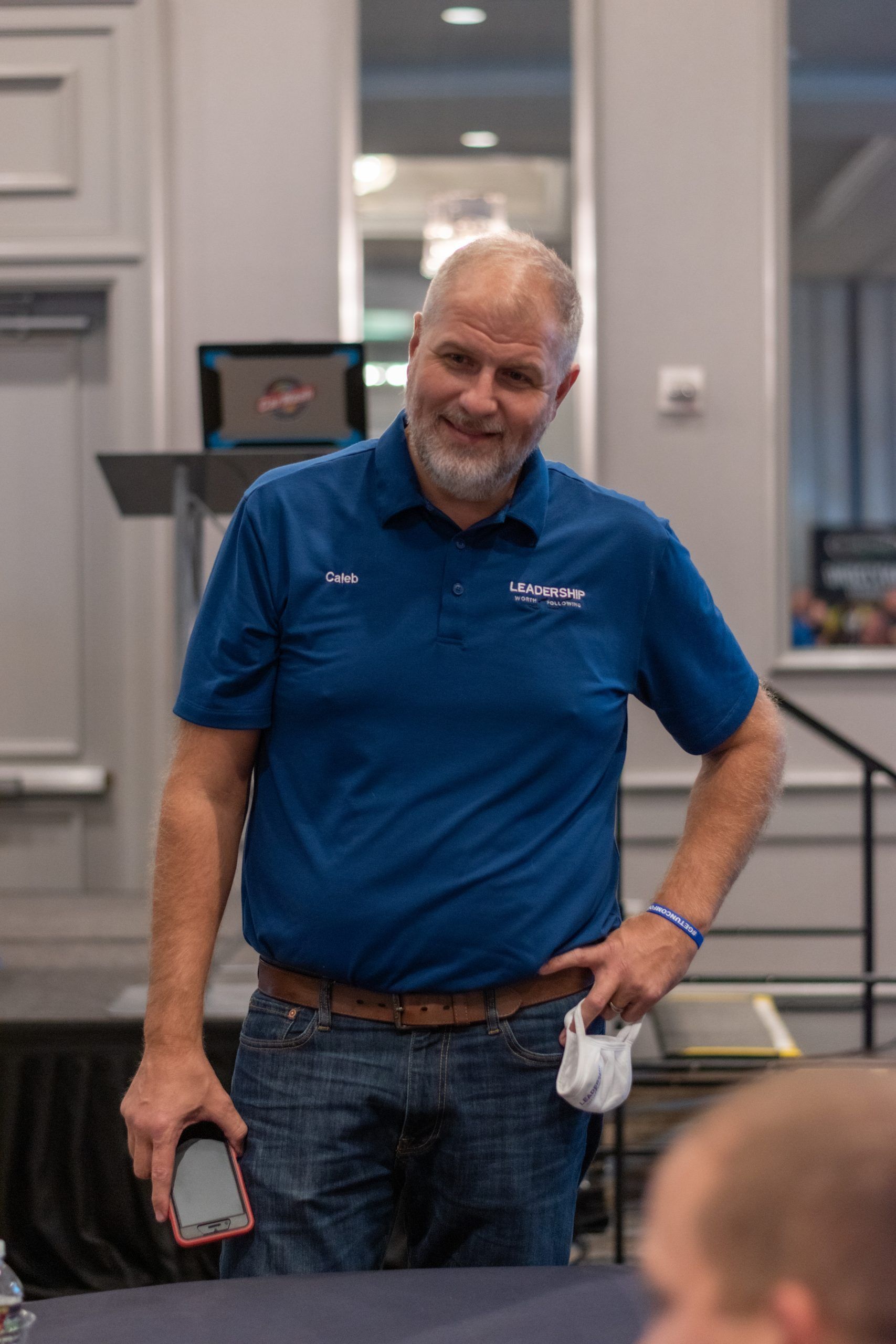 A man in a blue shirt is standing in a room holding a cell phone.