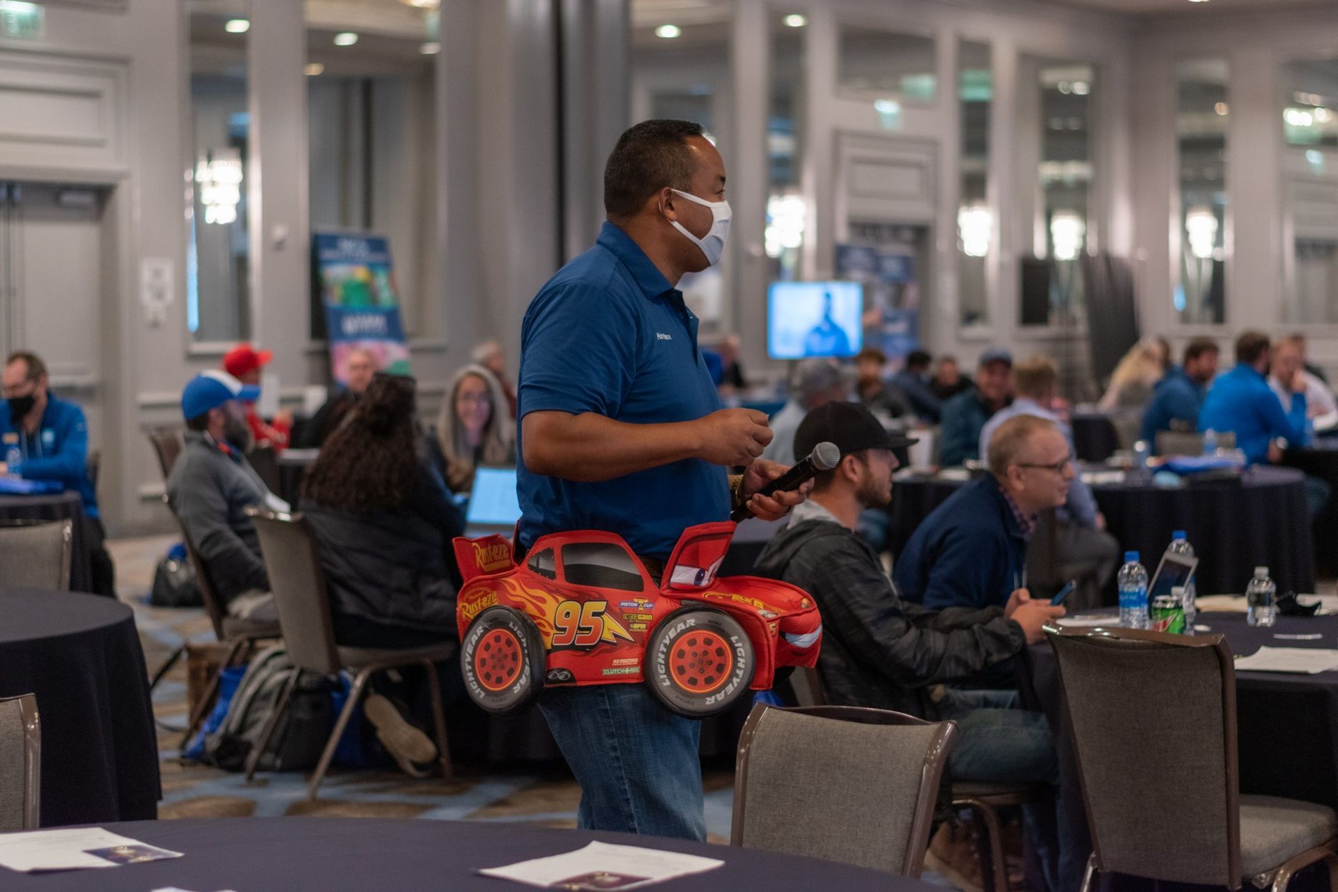 A man wearing a mask is holding a toy car in a conference room.