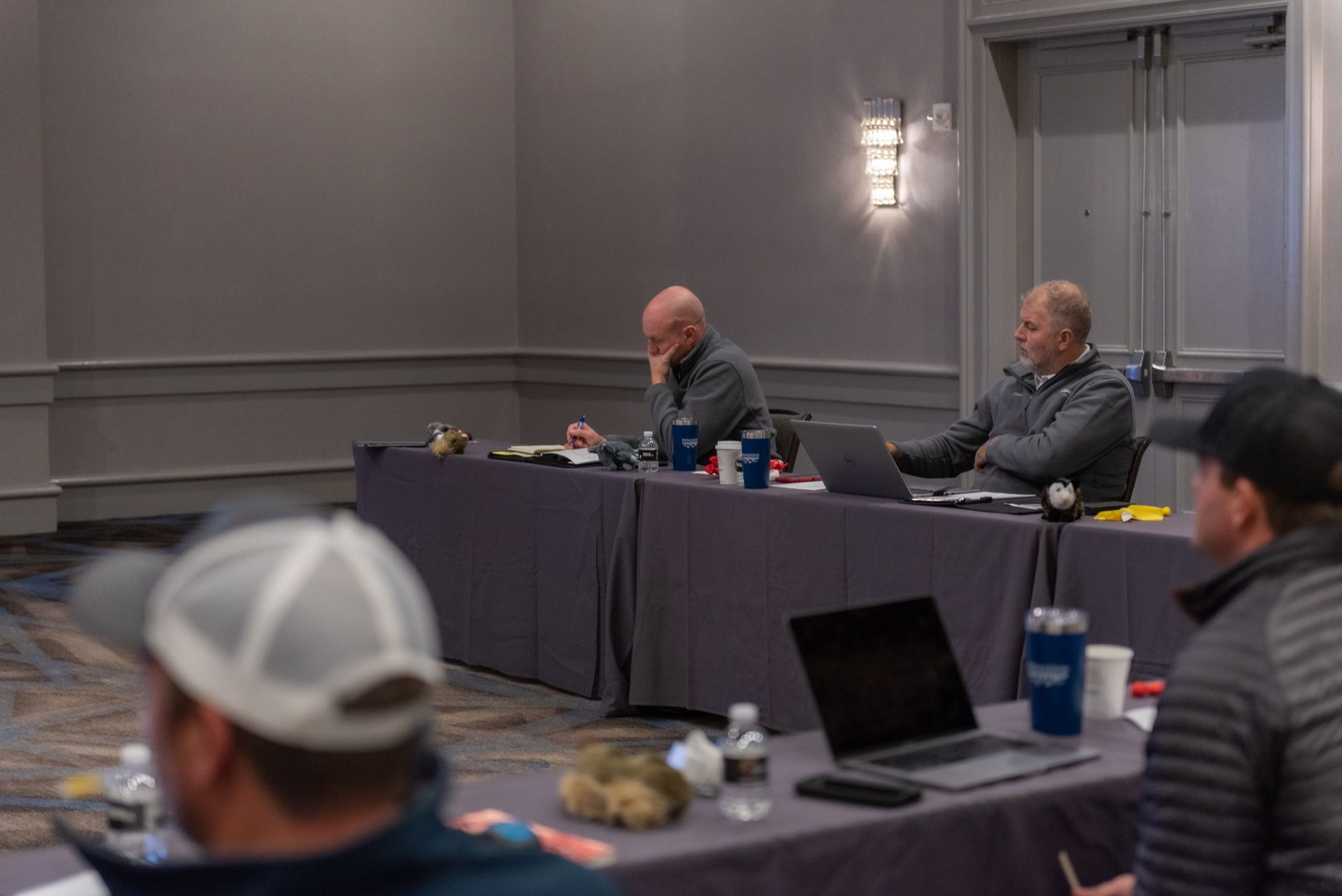 A group of men are sitting at long tables with laptops in a conference room.