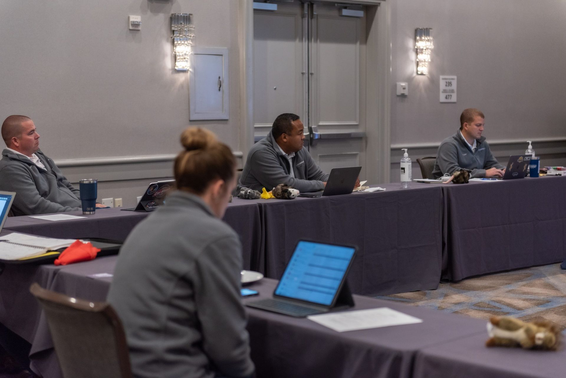 A group of people are sitting at long tables in a conference room.