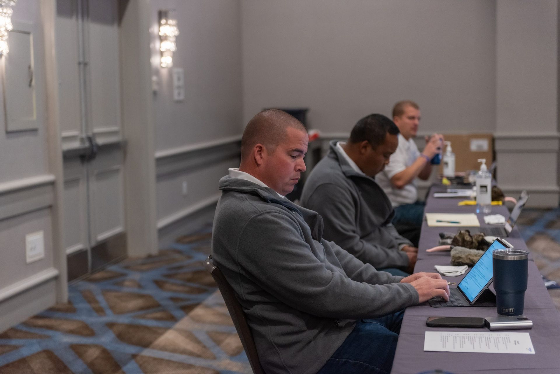 A group of men are sitting at a long table using laptops.