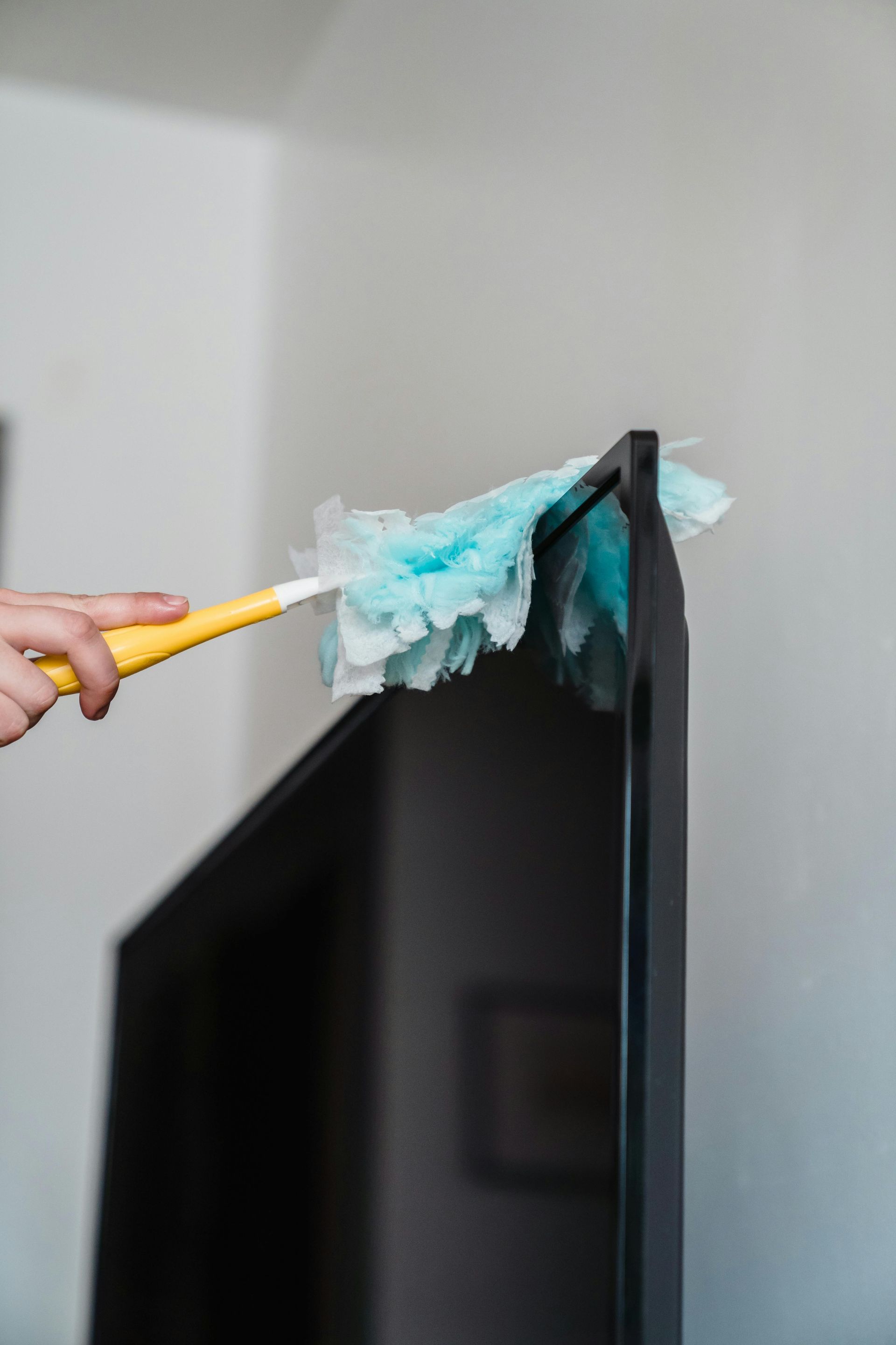 Hand dusting the top edge of a black TV with a yellow duster in a white room