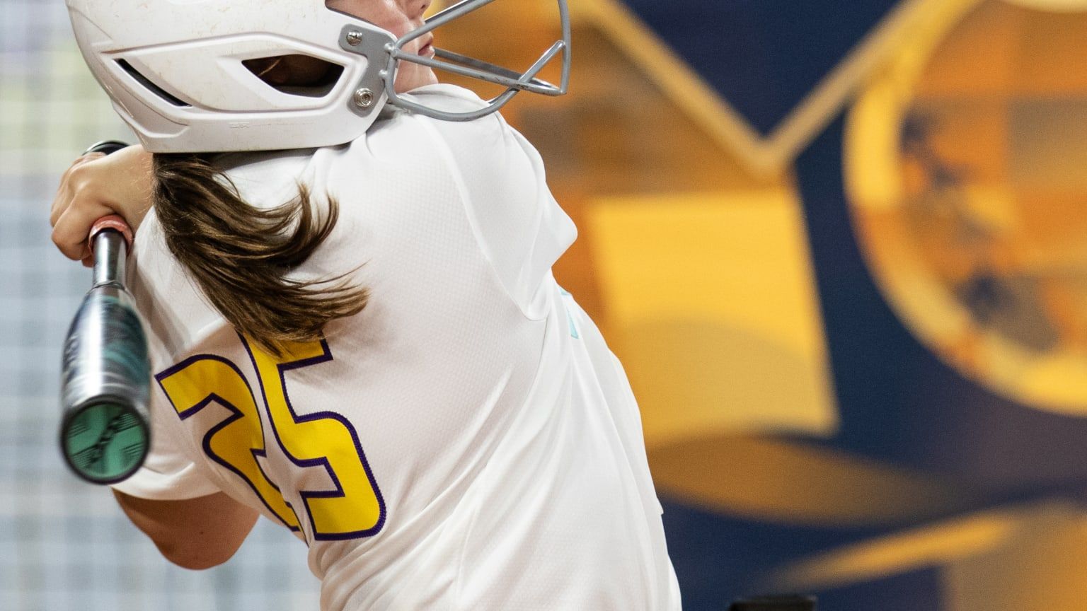 Softball player in white jersey with yellow number 35 swings a bat against a blurred stadium backdrop