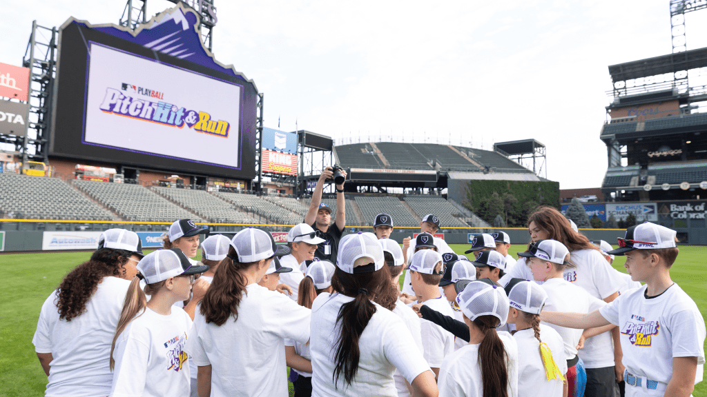 People in white shirts gather on a baseball field with a large scoreboard in the background.