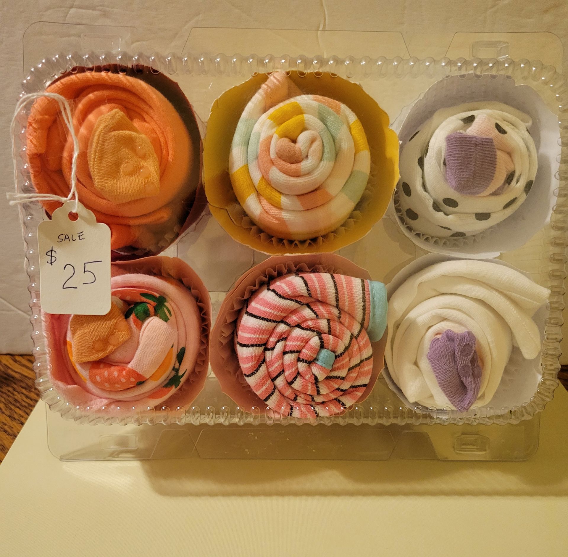 Six colorful frosted cupcakes in a clear plastic container, one with a $25 price tag.