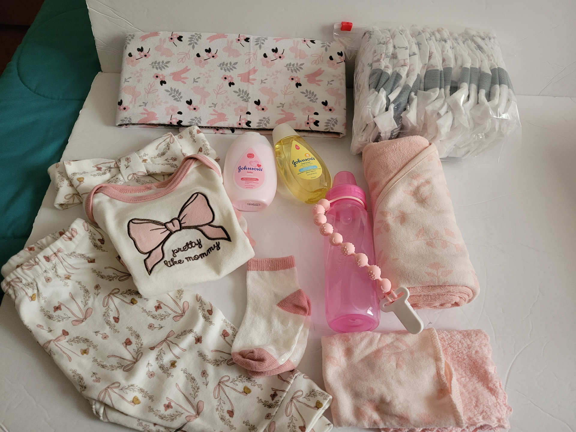 Assorted pink floral and pastel personal items laid out on a bed, including pouches, bottles, and a bottle carrier