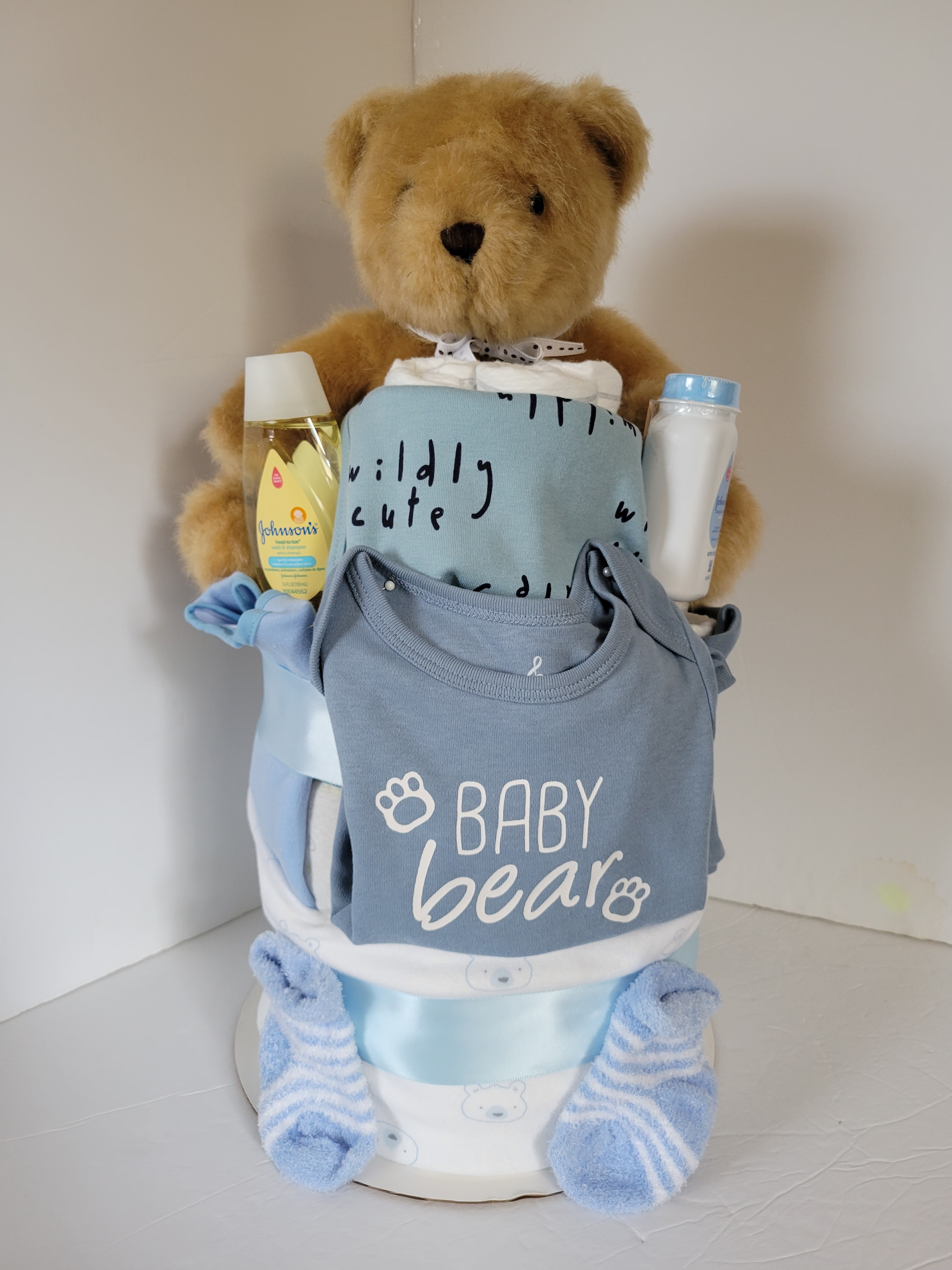 Teddy bear in a baby gift basket with a gray “Baby Bear” bib, cups, and blue booties