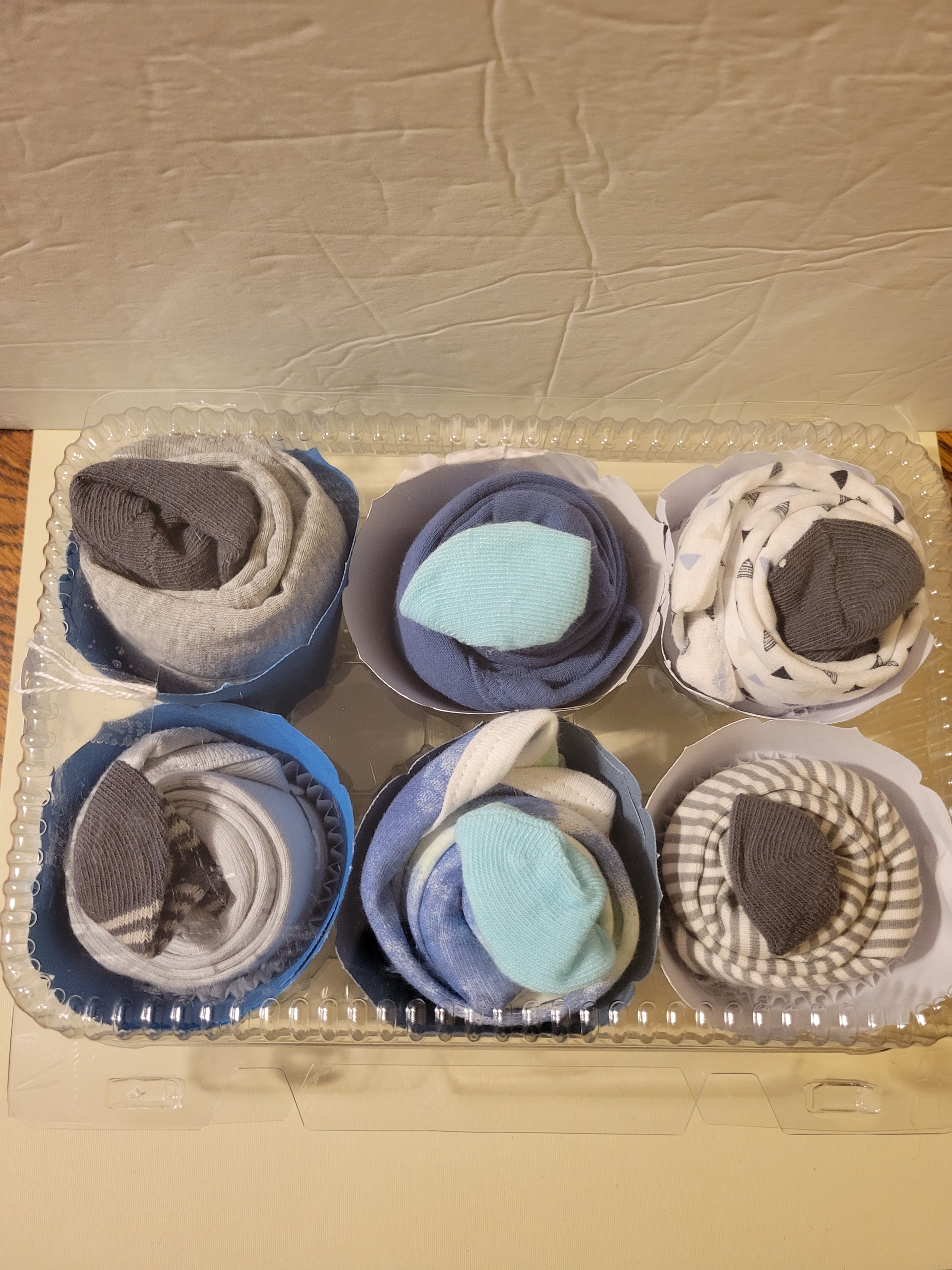 Six small folded fabric bundles in round blue, gray, and white containers on a cardboard surface.