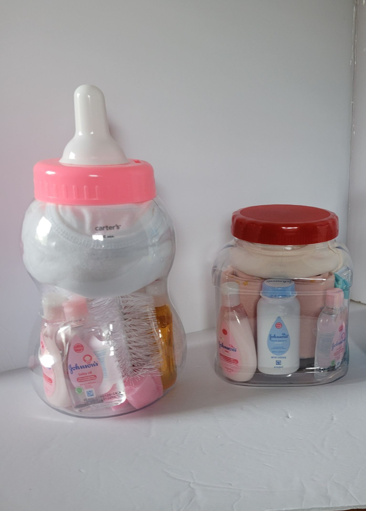 Two baby bottles and a jar of baby-care items on a wall shelf, one pink and one red-lidded.