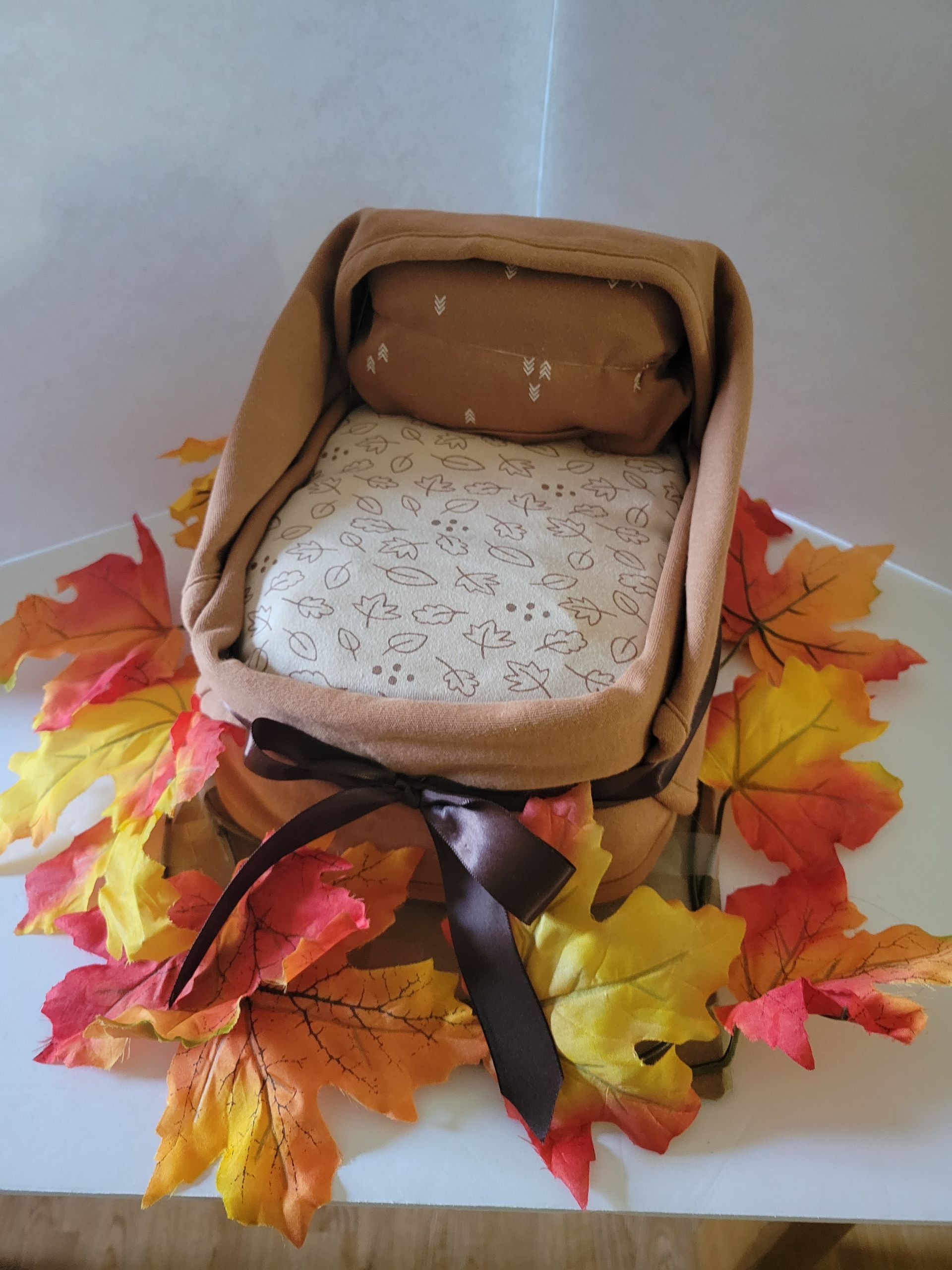 Miniature bed with tan pillows and white bedding, surrounded by autumn leaves and a brown ribbon.