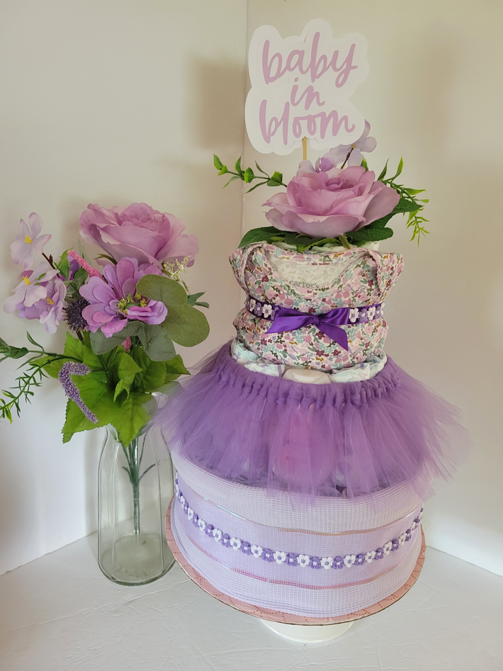 Purple baby shower diaper cake with flowers and “baby” topper beside a vase of blooms