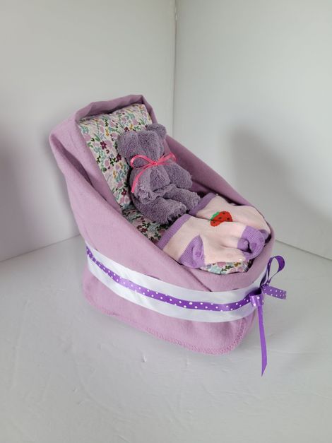 Purple fabric doll cradle with two plush toys and a patterned blanket, tied with a lavender ribbon