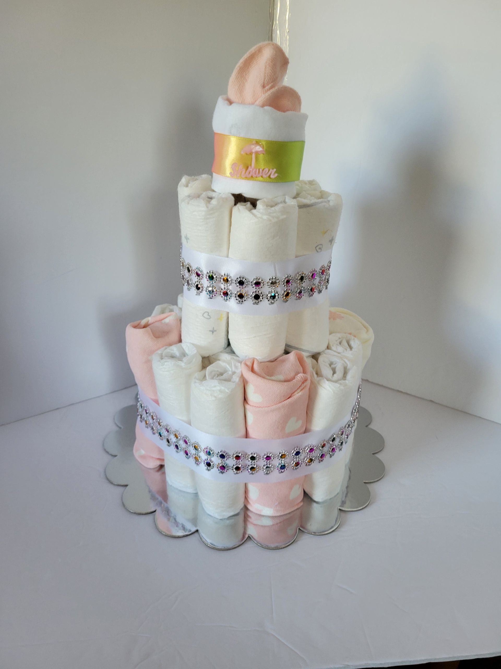 Tiered diaper cake with pastel pink and white rolled diapers, decorated with ribbon and topped with a baby bootie.