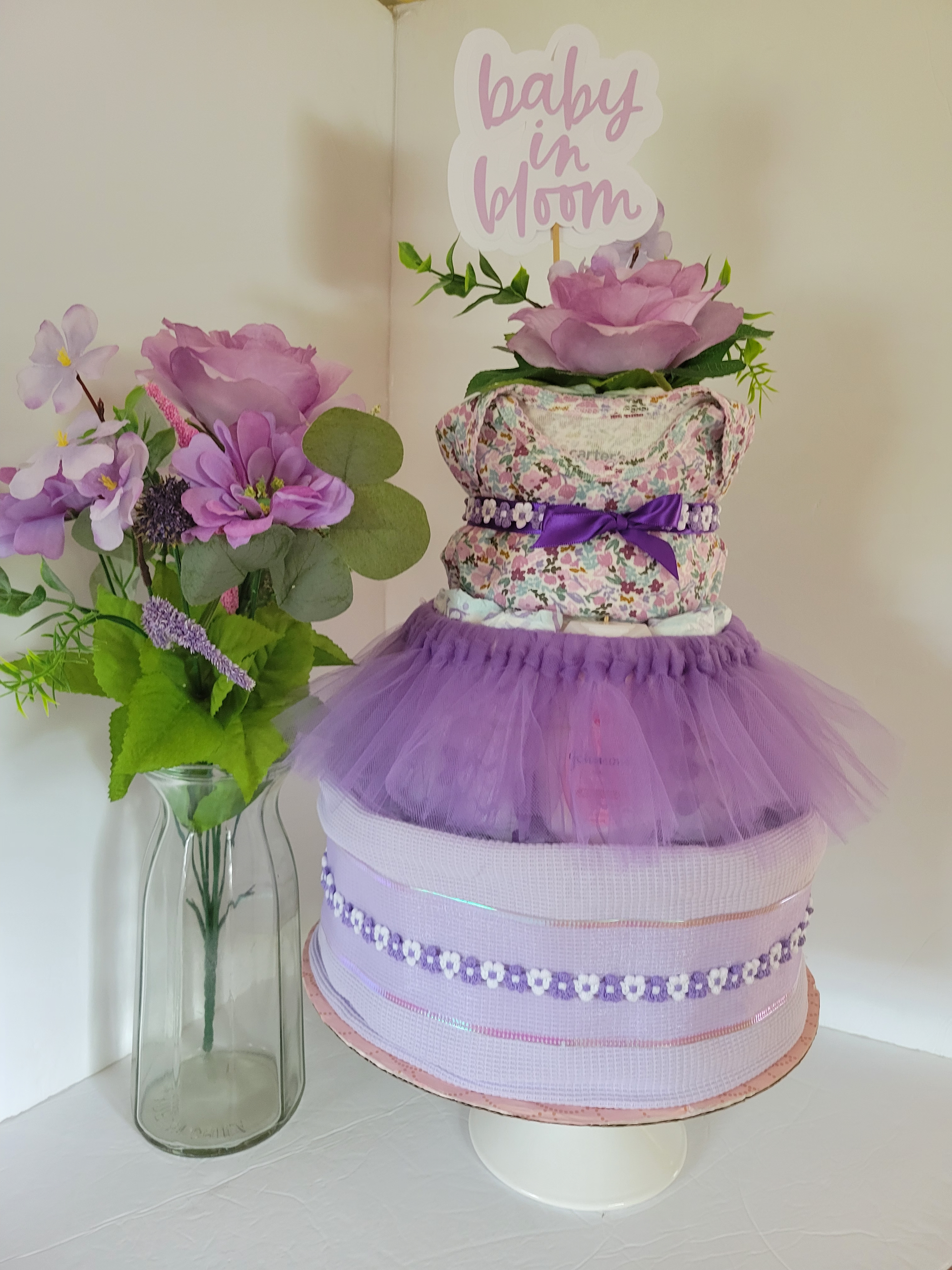 Purple tutu baby shower cake with floral topper beside matching flower arrangement in a vase