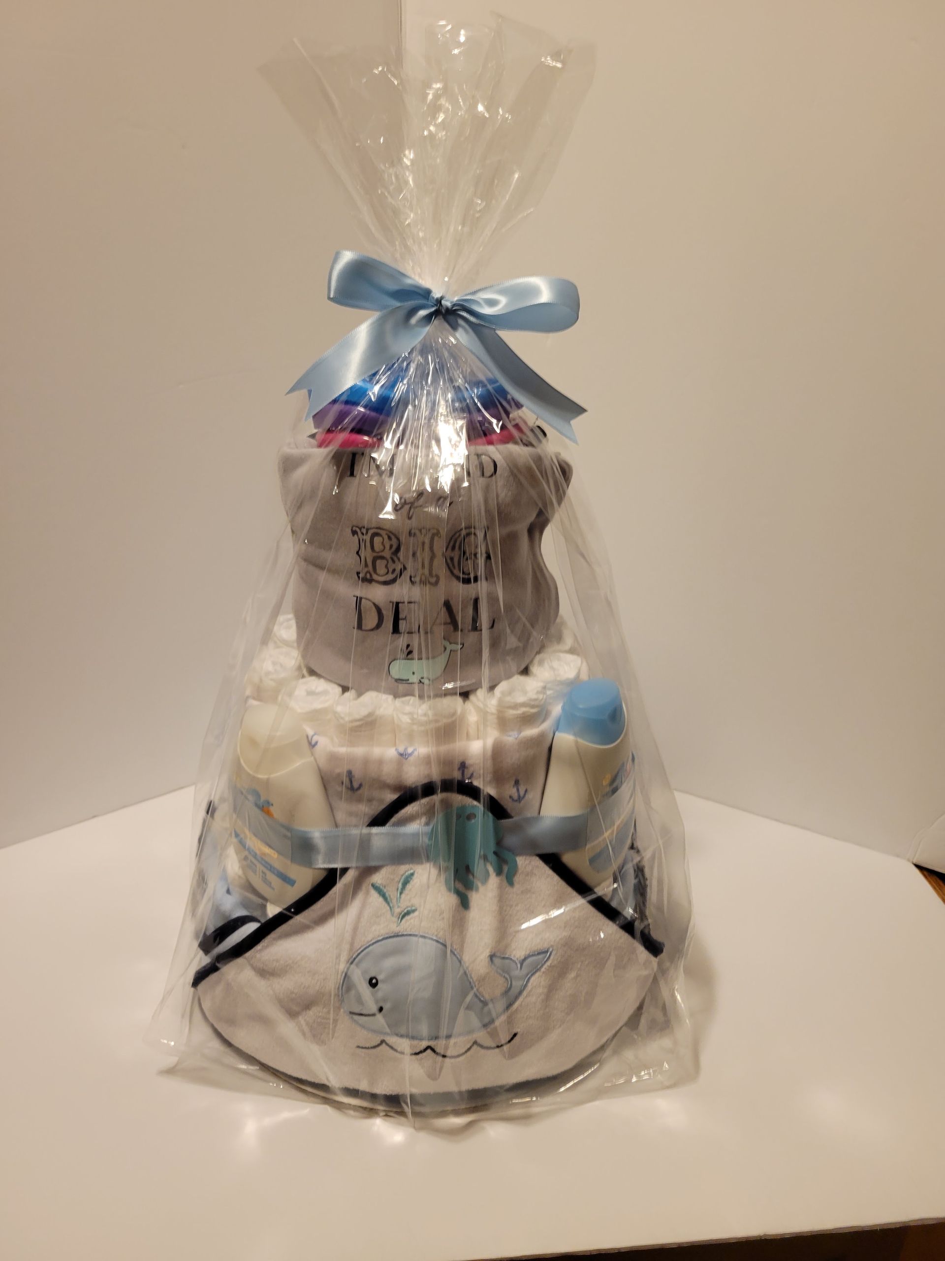Two-tier diaper cake with blue ribbon and baby-themed decorations, wrapped in clear cellophane.