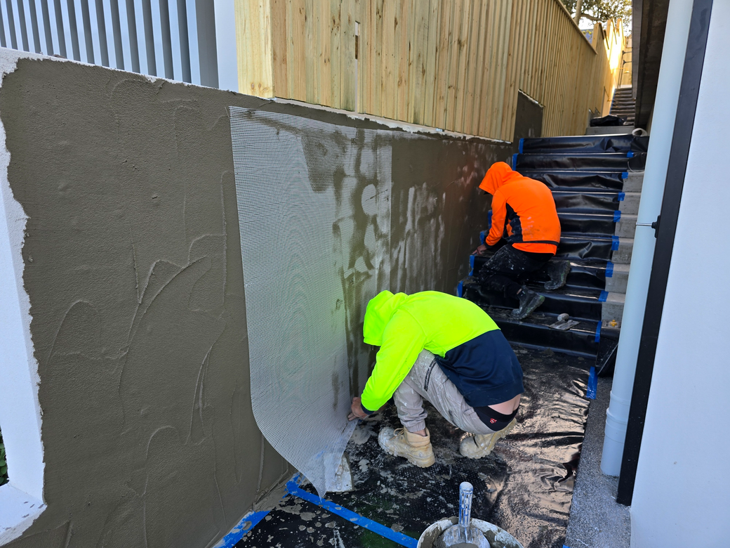 Two workers in bright jackets applying gray stucco to an exterior wall near outdoor stairs