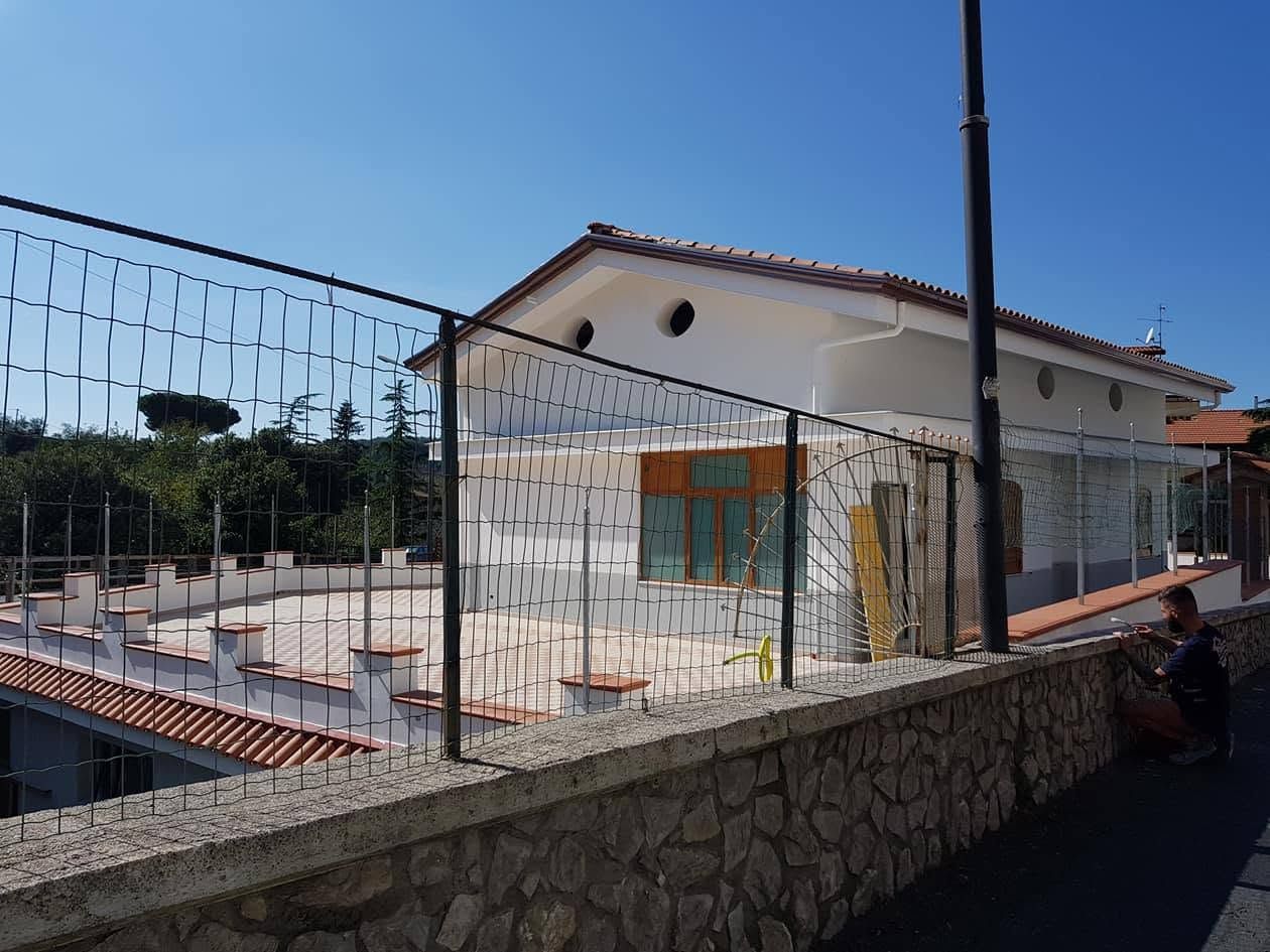 Terrazza esterna con muri in pietra e recinzione in filo metallico, e un edificio bianco sotto un cielo azzurro.