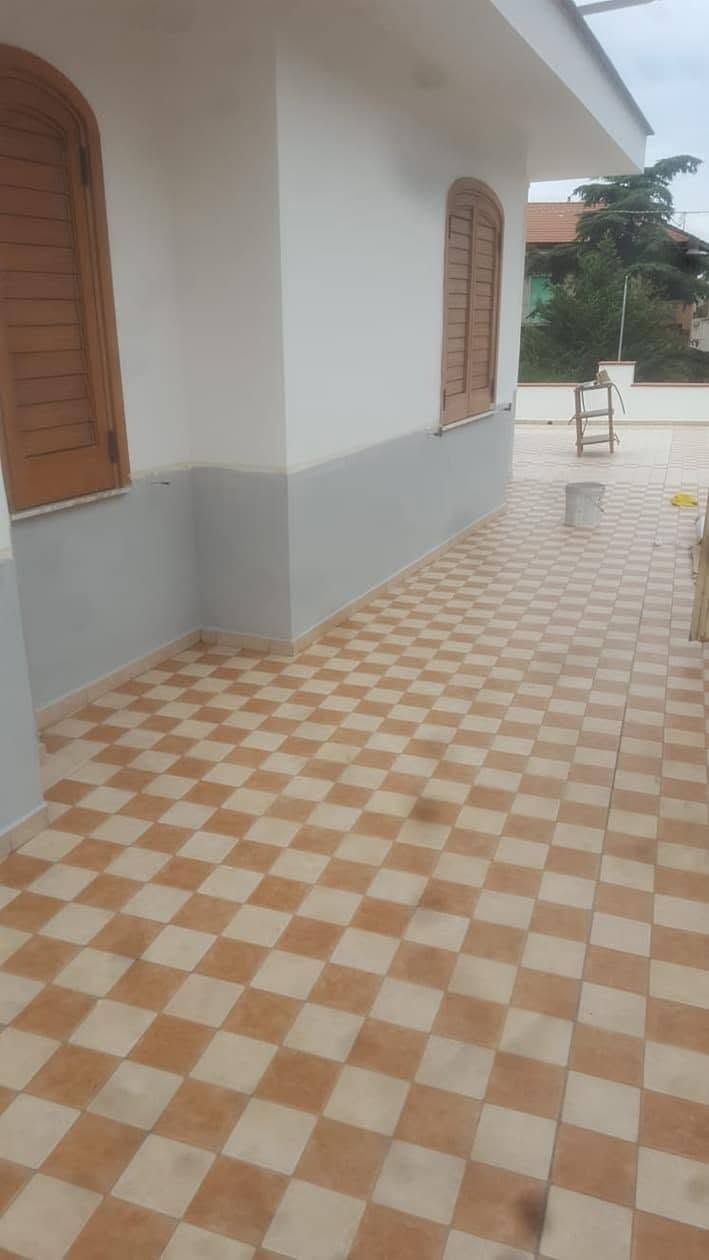Esterno della casa con portico piastrellato a scacchi beige e bianchi e finestre con persiane marroni.
