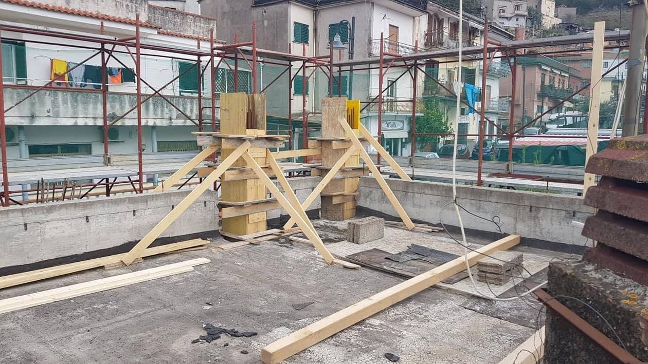 Cantiere edile con puntelli in legno a sostegno di una fondazione in cemento e impalcature sullo sfondo.