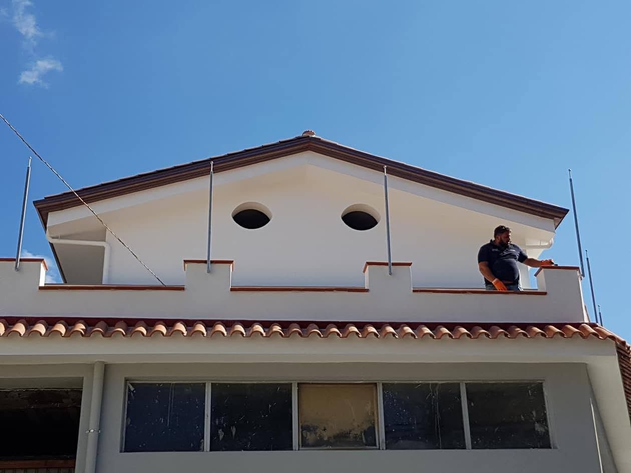 Persona al lavoro sul tetto di una casa bianca con finiture in terracotta, sotto un cielo azzurro.