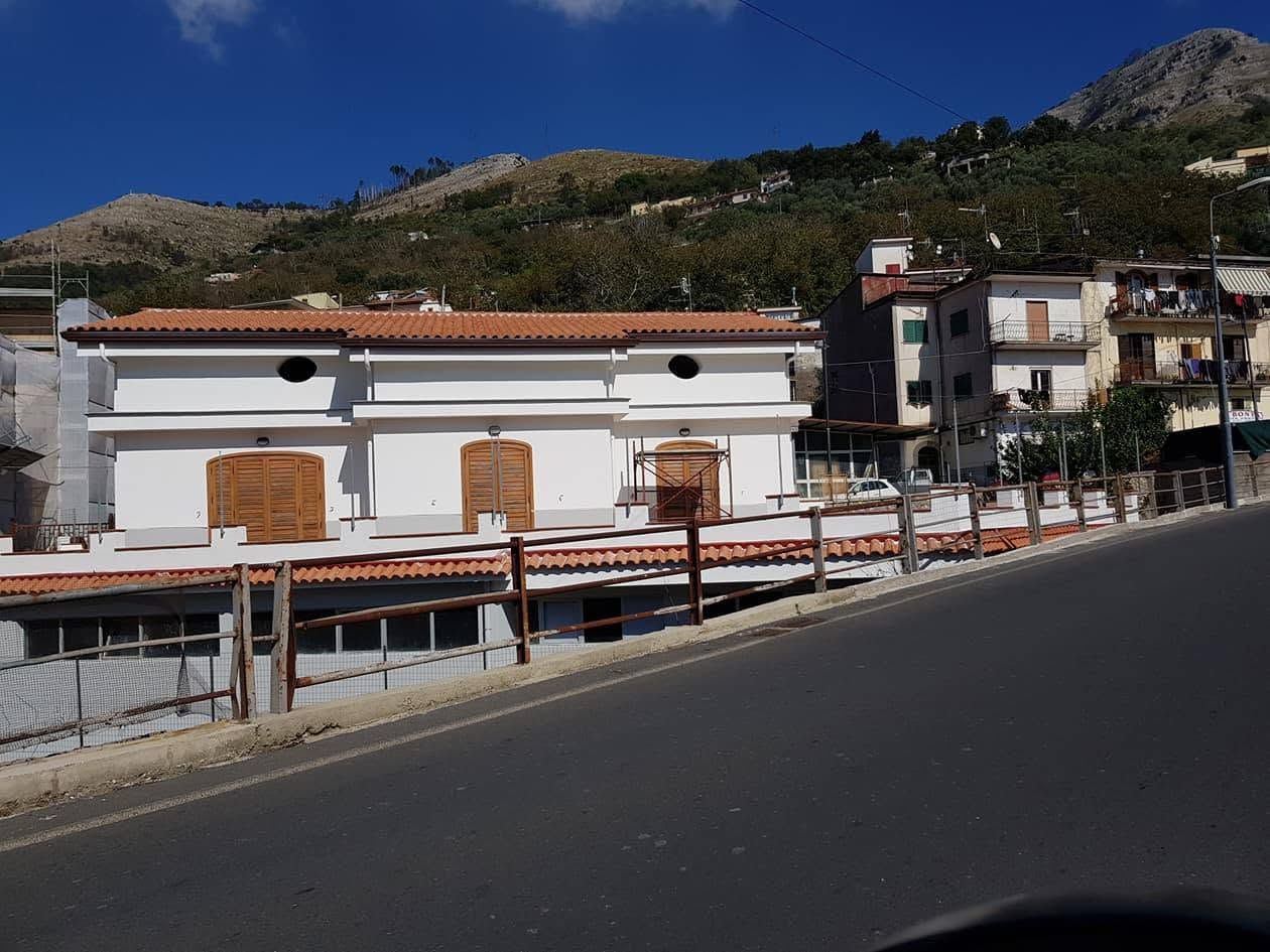 Edificio bianco con finestre sbarrate, situato accanto a una strada, con colline e cielo azzurro sullo sfondo.