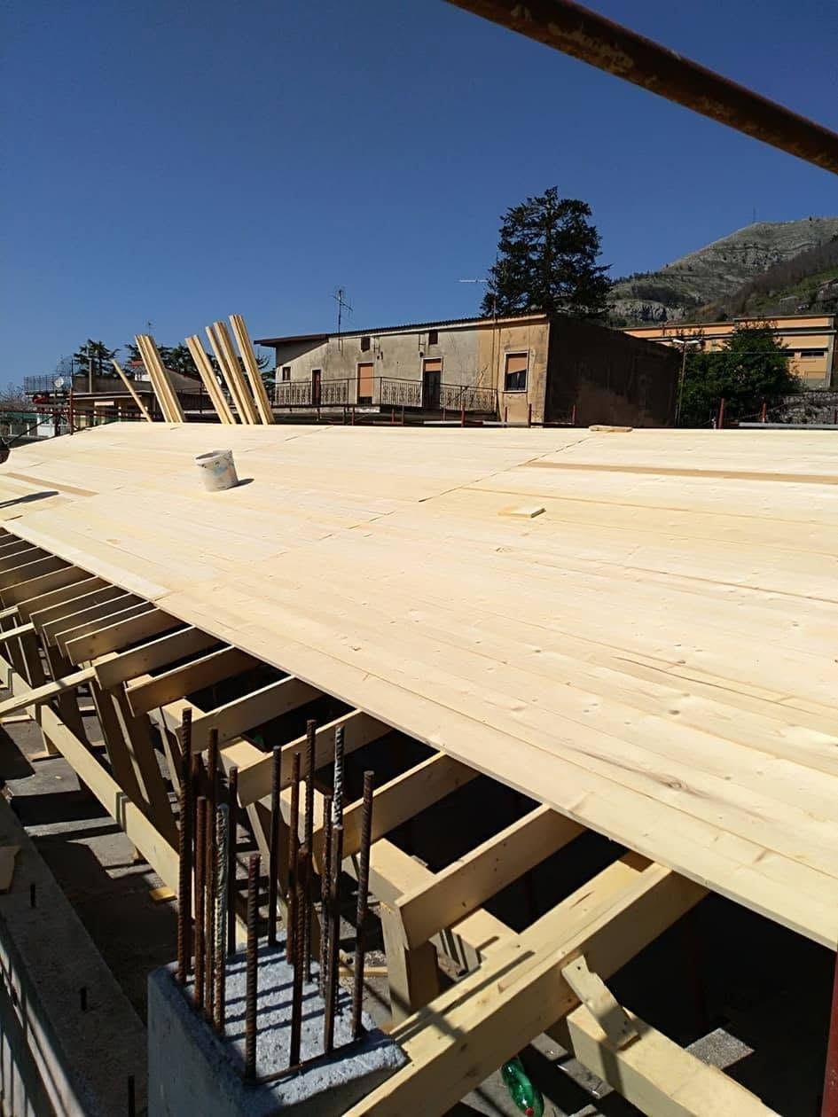 Cantiere edile con grandi casseforme curve in cemento, supporti in legno ed edifici sullo sfondo, sotto un cielo azzurro.