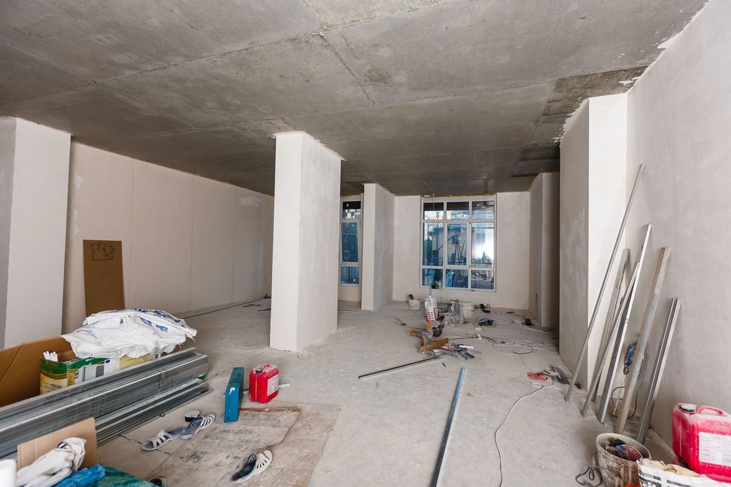 Interno di una stanza non rifinita con pareti bianche, soffitto in cemento a vista, attrezzi e materiali sparsi sul pavimento.