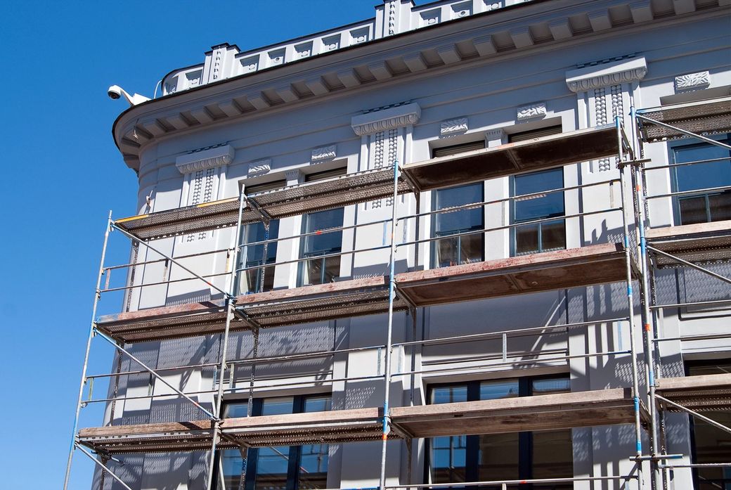 Facciata di un edificio storico in fase di ristrutturazione, con impalcature e reti di protezione, sullo sfondo di un cielo azzurro limpido.