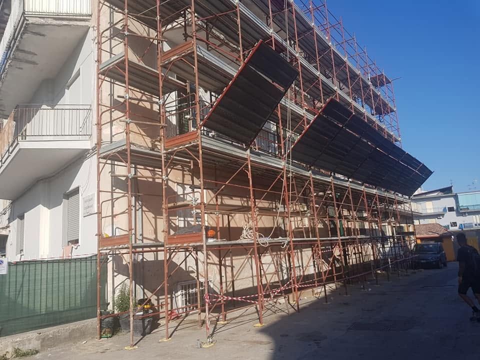 In un cantiere soleggiato, un'impalcatura copre la facciata di un edificio, con operai al lavoro nelle vicinanze.