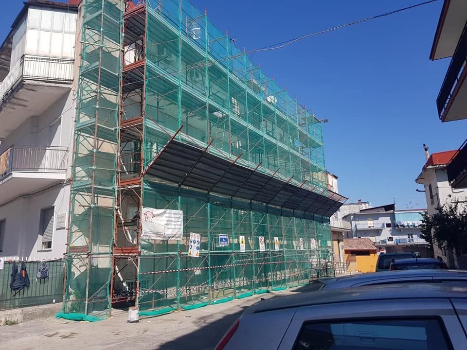 Edificio avvolto da impalcature verdi su una strada, con auto parcheggiate e cielo azzurro.