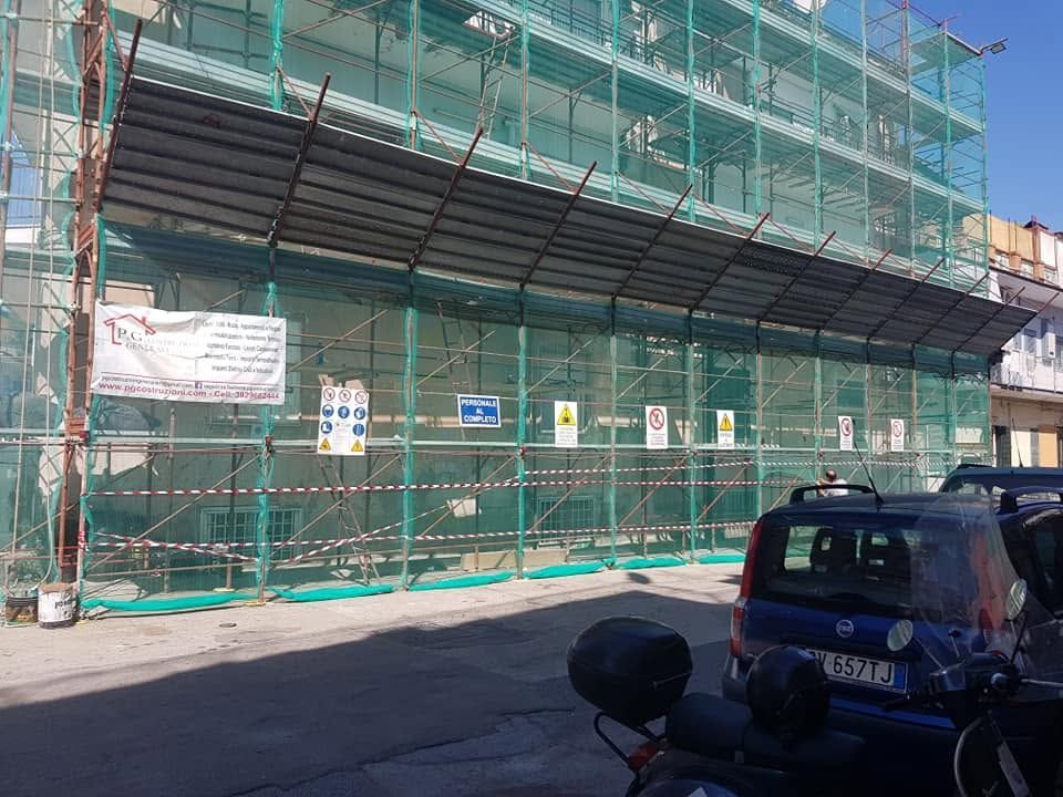 Cantiere edile con reti di sicurezza verdi e impalcature lungo una strada, con un furgone blu scuro parcheggiato davanti.