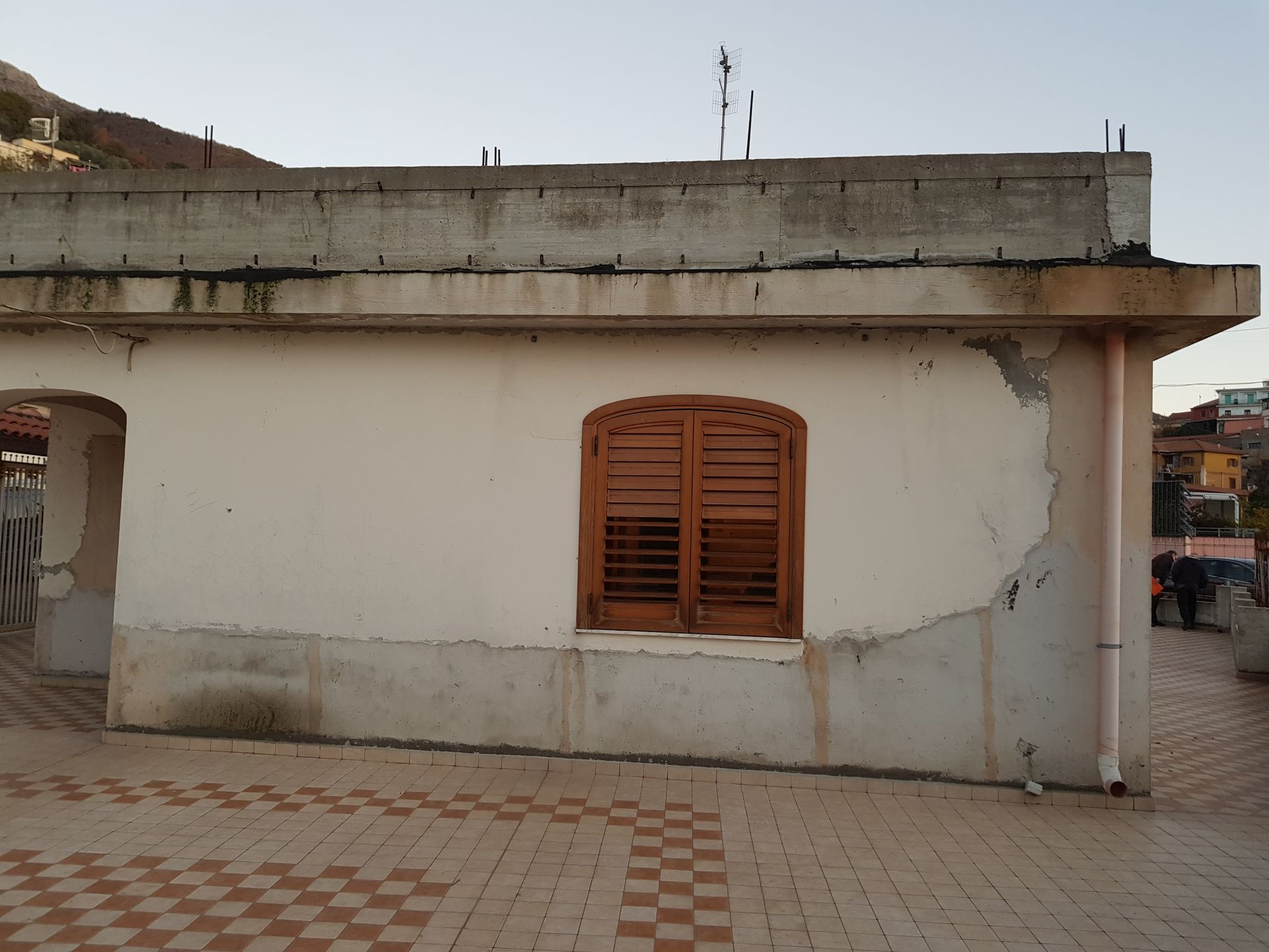 Piccolo edificio bianco con una finestra con persiane in legno e un cortile piastrellato sotto un tetto di cemento.