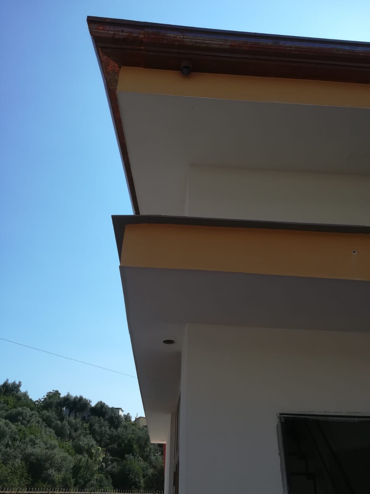 Primo piano del tetto e della grondaia di una casa, sullo sfondo di un cielo azzurro limpido, con alberi sottostanti.