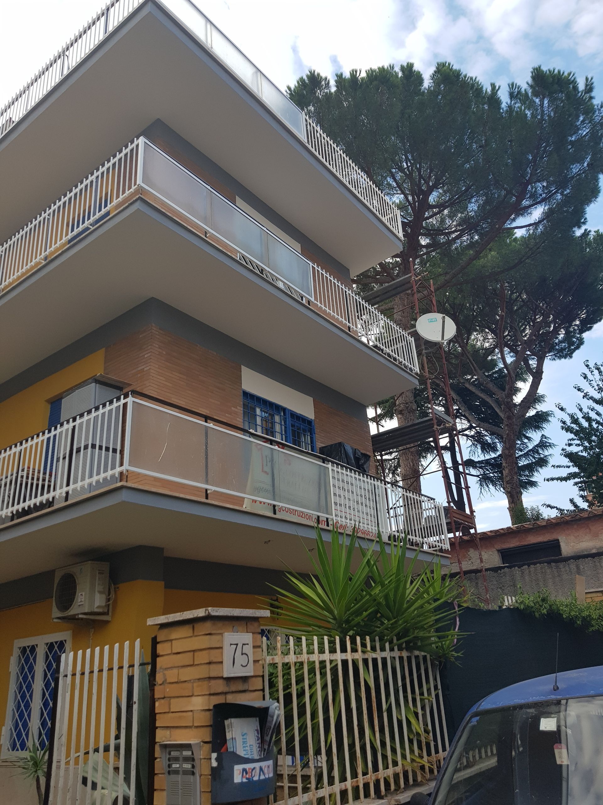 Edificio giallo a tre piani con balconi e un albero accanto a un'auto blu parcheggiata