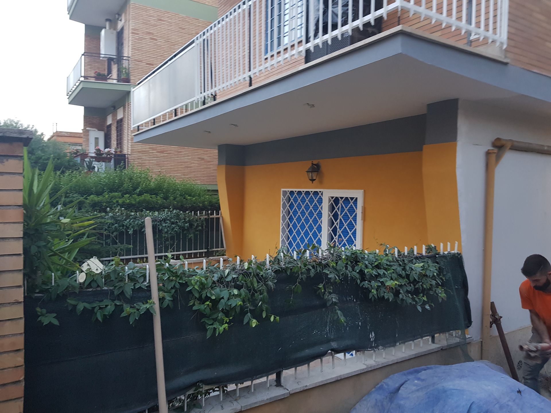 Casa con balcone e muro giallo dietro una siepe verde; persona in piedi a destra con un attrezzo