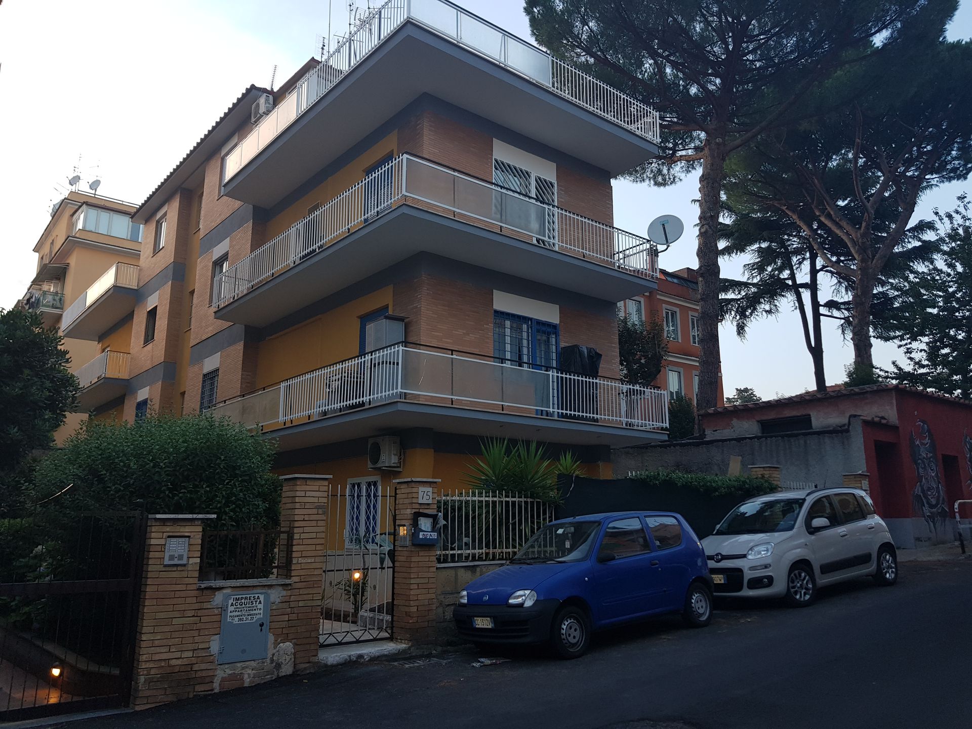 Edificio residenziale di tre piani con balconi, auto parcheggiate e alberi, situato in una strada residenziale.